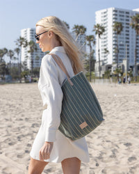 A woman with long blonde hair walks on a sandy beach under palm trees, wearing a white dress and sunglasses, carrying a Graf Lantz green canvas tote with leather straps. Tall buildings rise in the background beneath a clear blue sky.