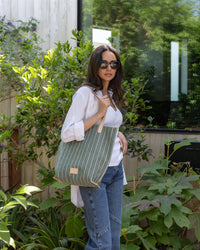 A woman stands outdoors near plants and a window, wearing sunglasses, a white shirt, and jeans while holding a large Graf Lantz green and white canvas tote with leather straps.