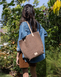 In a sunlit garden, a person with long, dark hair wears a denim shirt and brown shorts, carrying the striped Graf Lantz handbag with leather straps. Lush greenery surrounds them.