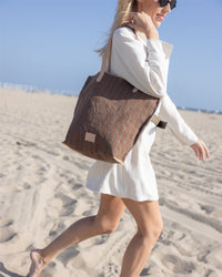 A woman in sunglasses and a white dress walks on a sandy beach with a Graf Lantz canvas tote with leather straps over her shoulder under a clear blue sky.