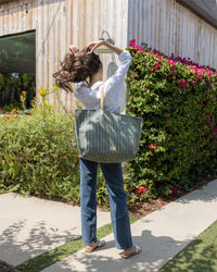 A woman stands outdoors with her back to the camera, holding her long brown hair. She wears a white shirt, blue jeans, sandals, and carries a versatile Graf Lantz carryall with leather handles amid greenery and pink flowers.