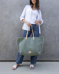 A woman in a white shirt, white tank top, blue jeans, and white sandals poses by a gray wall, holding a large green striped Graf Lantz canvas boat bag with tan leather handles.