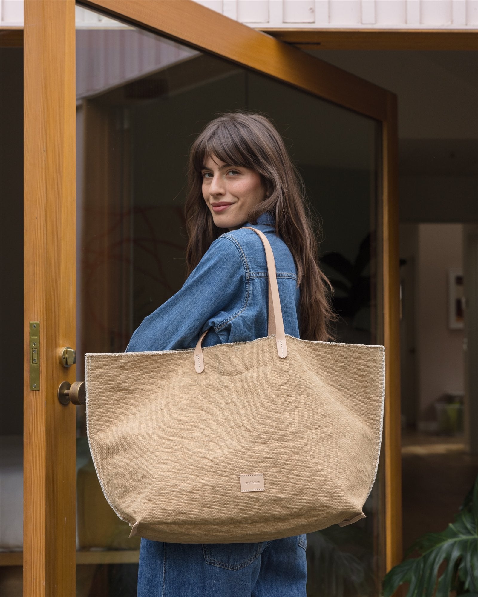 A person with long hair stands in a doorway, wearing denim and shouldering a Graf Lantz Hana Canvas Boat Bag with genuine leather handles, against the backdrop of an open wooden door and part of a spacious interior.
