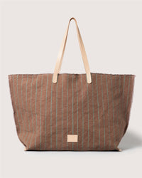 A large brown canvas boat bag by Graf Lantz features thin green vertical stripes, beige double handles, frayed edges for a casual look, and a small beige patch near the bottom center.