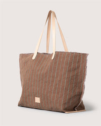 A spacious brown canvas boat bag by Graf Lantz features light green vertical pinstripes, beige leather handles, and a small front patch—this versatile carryall is shown against a plain light background.