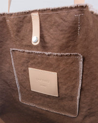 Close-up of a brown canvas Graf Lantz boat bag tote with a front pocket featuring raw edges and a beige leather patch with embossed text. Versatile carryall has beige leather handles secured by metal studs.