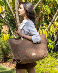 A woman with long dark hair, in a white shirt and brown skirt, walks outdoors surrounded by greenery, carrying a Graf Lantz versatile carryall with leather handles over her shoulder.