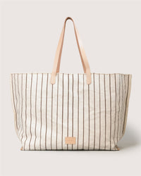 A large beige Graf Lantz Hana Boat Bag canvas tote with vertical thin brown stripes, tan leather handles, and a small rectangular tan leather patch on the front, shown against a light background.