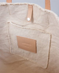 Close-up of the inside of a beige canvas Hana Boat Bag by Graf Lantz, showing a stitched pocket with a rectangular tan leather label that reads Graf Lantz and a leather strap secured with a metal rivet.