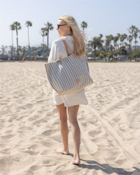 A woman with long blonde hair and sunglasses walks barefoot on the beach, carrying a large Graf Lantz Hana Boat Bag with leather handles. Palm trees and a clear sky are in the background.