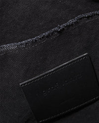 Close-up of textured black fabric with visible stitching, exuding the refined elegance of Graf Lantzs Hana Hobo. A black label partially wraps around the edge, bearing subtle engraved text in a minimalist style similar to the exclusive Hana Bag Collection.
