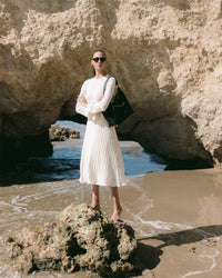 On a rocky beach with a grand rock arch behind her, a woman in a flowing white dress confidently wears sunglasses while holding a chic black shoulder bag from the Graf Lantz Hana Bag Collection, as gentle waves wash ashore.