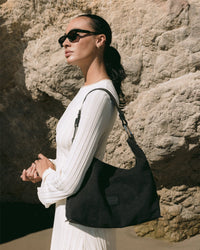 A woman in a white dress and sunglasses stands against a rocky background, holding a sleek black shoulder bag from the Graf Lantz Hana Collection. Her dark hair is slicked back as she gazes into the distance, in an outdoor setting with natural lighting.