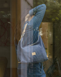 Inside, a long-haired person wearing a denim jacket showcases the Hana Hobo by Graf Lantz. Reflections in the glass window reveal vibrant outdoor greenery and stylish indoor decor.