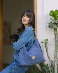 A person with long hair leans against a wall, smiling slightly. They wear blue denim jeans and jacket, effortlessly carrying a large Hana Hobo shoulder bag by Graf Lantz. A cactus is in the foreground, while a wooden doorframe behind them adds to the scenes charming simplicity.