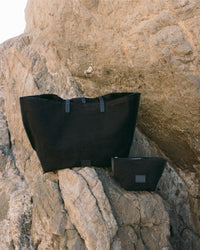 On rugged rocks, two black fabric bags—one large and one small—from Graf Lantz showcase their artisanal Hana Pouch design, featuring minimalist aesthetics with subtle details. Natural stone formations provide a striking backdrop.