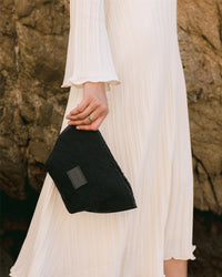 A person in a long white dress holds a Graf Lantz artisanal black clutch against a textured rock background. The dress features subtle stripes and flared sleeves, showcasing fine craftsmanship. Only the lower half of the persons body and hand are visible.