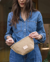 A person in a blue denim shirt holds a Graf Lantz Hana Canvas Pouch with both hands. The pouch, featuring a small rectangular label and artisanal fraying, is showcased against a softly lit indoor background that highlights its crafted charm.