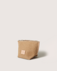 A small, light brown Graf Lantz Hana Canvas Pouch with a rectangular beige label on the front stands upright on a plain, off-white background, highlighting its elegant simplicity.