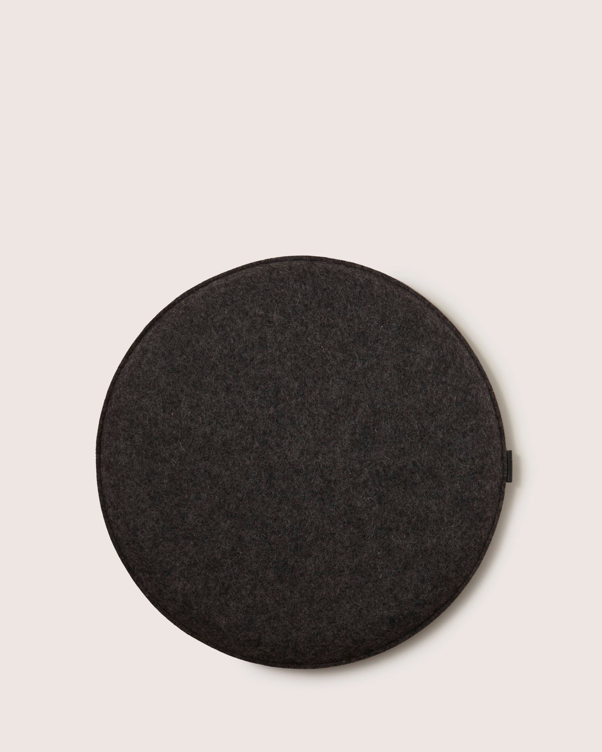 The graf lantz round Zabuton seat cushion in dark gray felt is made from soft Merino wool. It features a minimalist design, making it ideal for meditation or relaxation, and it rests against an off-white background.