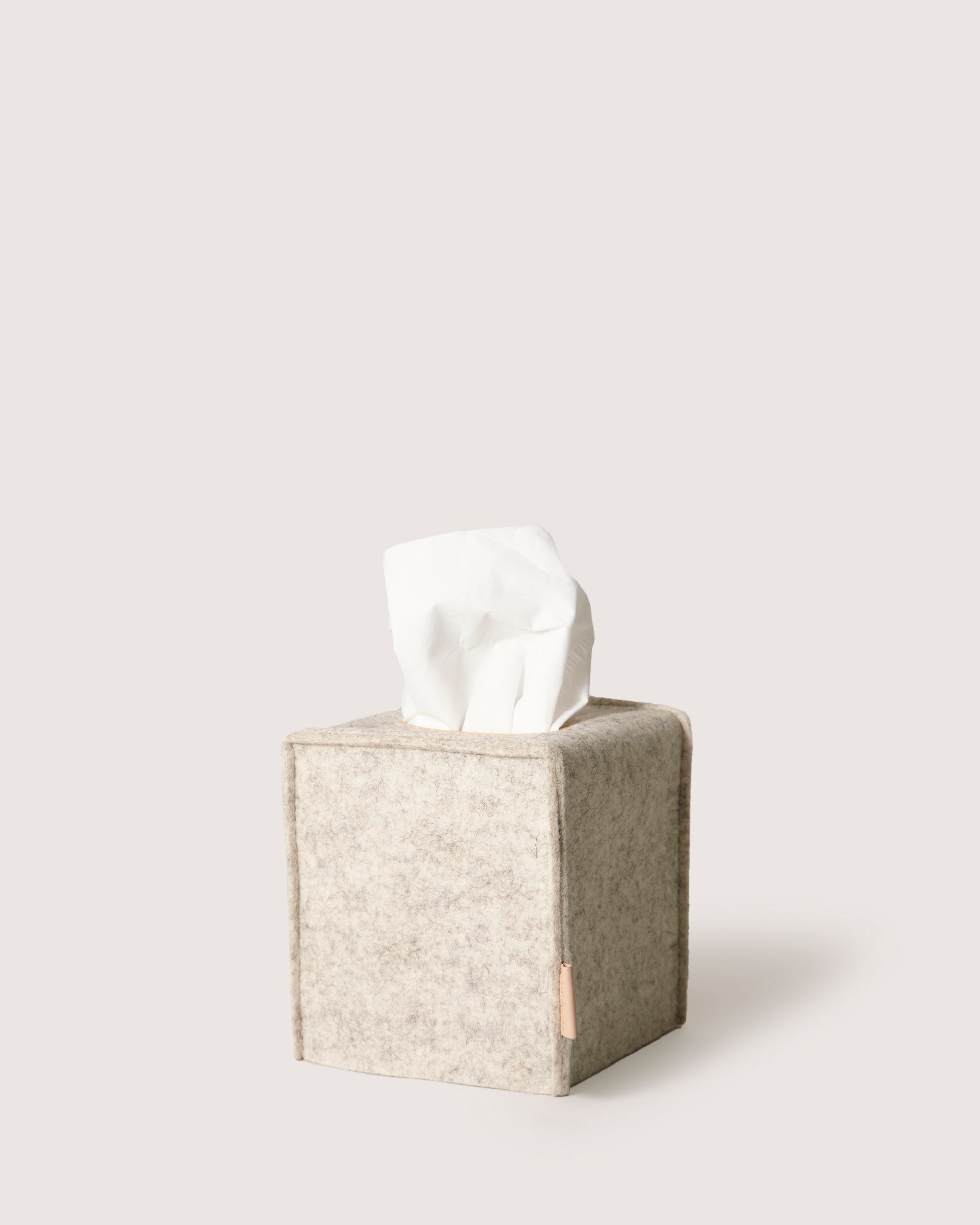 A sustainable beige felt tissue box cover by graf lantz, made from Merino wool, showcases a single white tissue gracefully emerging from the top on a light gray background.