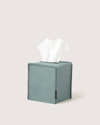 A light gray tissue protrudes from a square, light blue Graf Lantz Merino wool tissue box cover against a plain off-white background—an elegant touch for minimalist home decor.