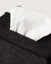 A close-up shows a white tissue partly pulled from a graf lantz dark, textured box with a minimalist silhouette akin to premium Merino wool. The plain, neutral background enhances the elegant simplicity of its design.