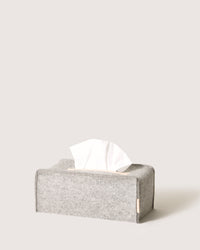 A gray felt tissue box cover by graf lantz, crafted from sustainable Merino wool with a wooden button accent. Designed to hold a tissue box with one tissue peeking out, set against a simple light background.