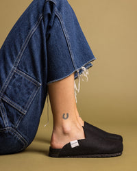 A person in frayed blue jeans sits on the floor, revealing a horseshoe tattoo on their lower leg while wearing black slip-on shoes crafted from Merino wool felt with a visible fabric tag by graf lantz. The background is plain olive.