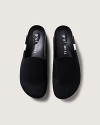 A pair of black slip-on slippers from graf lantz, crafted from soft Merino wool felt with the brand name on the insole. They feature white and black striped side tabs against a light gray background.