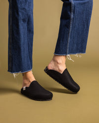 A person in frayed blue jeans steps on a mustard-colored floor, wearing graf lantz black slip-on shoes featuring textured recycled rubber soles.