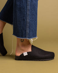 Wearing graf lantz black slip-on shoes with recycled rubber soles and blue frayed jeans against a muted yellow backdrop. A small label is on the shoe, and a tattoo peeks above the heel.