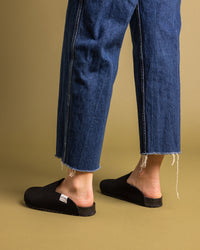 Sporting graf lantzs black slip-on shoes with recycled rubber soles and a small white tag on the side, a person stands on a tan background. Theyre wearing frayed-hem blue jeans featuring noticeable side stitching.