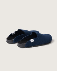 A pair of graf lantz dark blue slip-on slippers features a minimalist design on a light background, with black soles, a small white side tag, insoles made from recycled materials, and a cork footbed for added comfort.