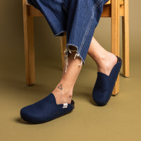 A person sits on a wooden chair wearing graf lantz dark blue felt slippers with recycled rubber soles, frayed hem blue jeans, and a small triangle tattoo on their right ankle. The warm beige background enhances the cozy ambiance.