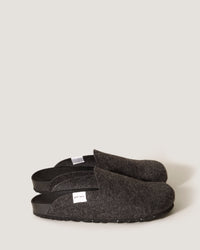 A pair of dark gray graf lantz Merino wool felt slippers with black soles rests on a light gray background. Featuring an open-heel design and a small white side label, these slippers showcase sustainable footwears blend of style and comfort.