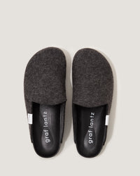 A pair of dark gray Merino wool felt slippers from graf lantz is viewed from above. They feature black leather insoles with white text and a small white tag on each slipper. The design is simple and sustainable with a light gray background.