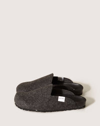 A pair of dark gray slip-on slippers by graf lantz, made from Merino wool felt with black rubber soles and a small white side tag, set against a plain light background. These stylish footwear pieces are both sustainable and eco-friendly.