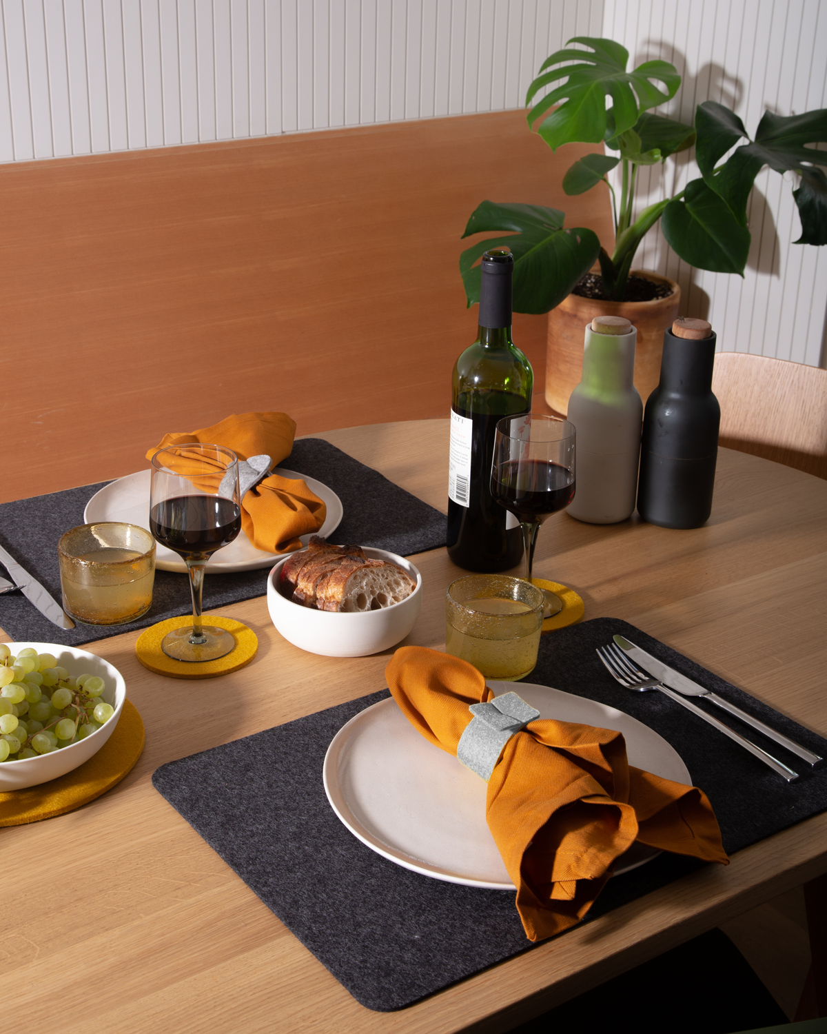 A neatly arranged dining table for two showcases white plates on graf lantz Merino wool placemats, mustard napkins, a bowl of grapes, bread loaf, two wine glasses with a red wine bottle, two water bottles, and a potted plant in the back.