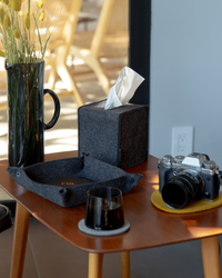 A cozy indoor scene features a camera on a yellow coaster, a graf lantz felt tissue box cover, and a sleek black vase with flowers on a wooden table. Nearby are a folded Merino wool basket and dark glass cup. Warm light filters through, enhancing the inviting home décor ambiance.