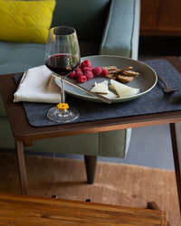 A glass of red wine is on a wooden table with a premium Merino wool placemat from graf lantz, beside a plate of raspberries, cheese, and bread. A folded napkin and knife are nearby, while in the background, a green sofa with a yellow pillow completes this designer-inspired look.