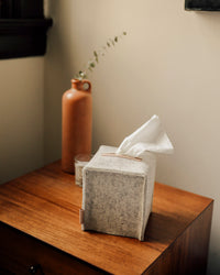 A graf lantz sustainable gray fabric tissue box cover sits on a wooden table, with a tissue poised for use. In the background, a brown ceramic vase with greenery and a small candle complement the cozy minimalist vibe.