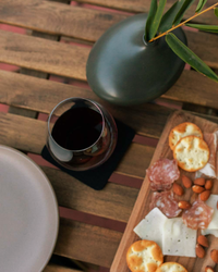 A wooden table features a charcuterie board with crackers, salami, cheese, and almonds. A glass of red wine sits on a graf lantz Merino wool coaster. A minimalist dark vase with green leaves completes the elegant scene.