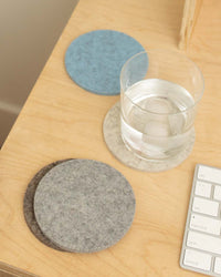 A glass of water sits on a light gray coaster made of Merino wool from graf lantz, positioned on a wooden desk. Nearby, two more coasters in blue and dark gray German Merino wool from graf lantz are stacked neatly. In the bottom right corner, the edge of a white keyboard is visible.