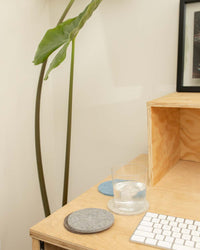 A minimalist workspace showcases a wooden desk with a glass of water and three round graf lantz coasters made from German Merino wool in gray and blue. A large-leafed plant and a framed picture accentuate the background, while a keyboard is partially visible on the desk.
