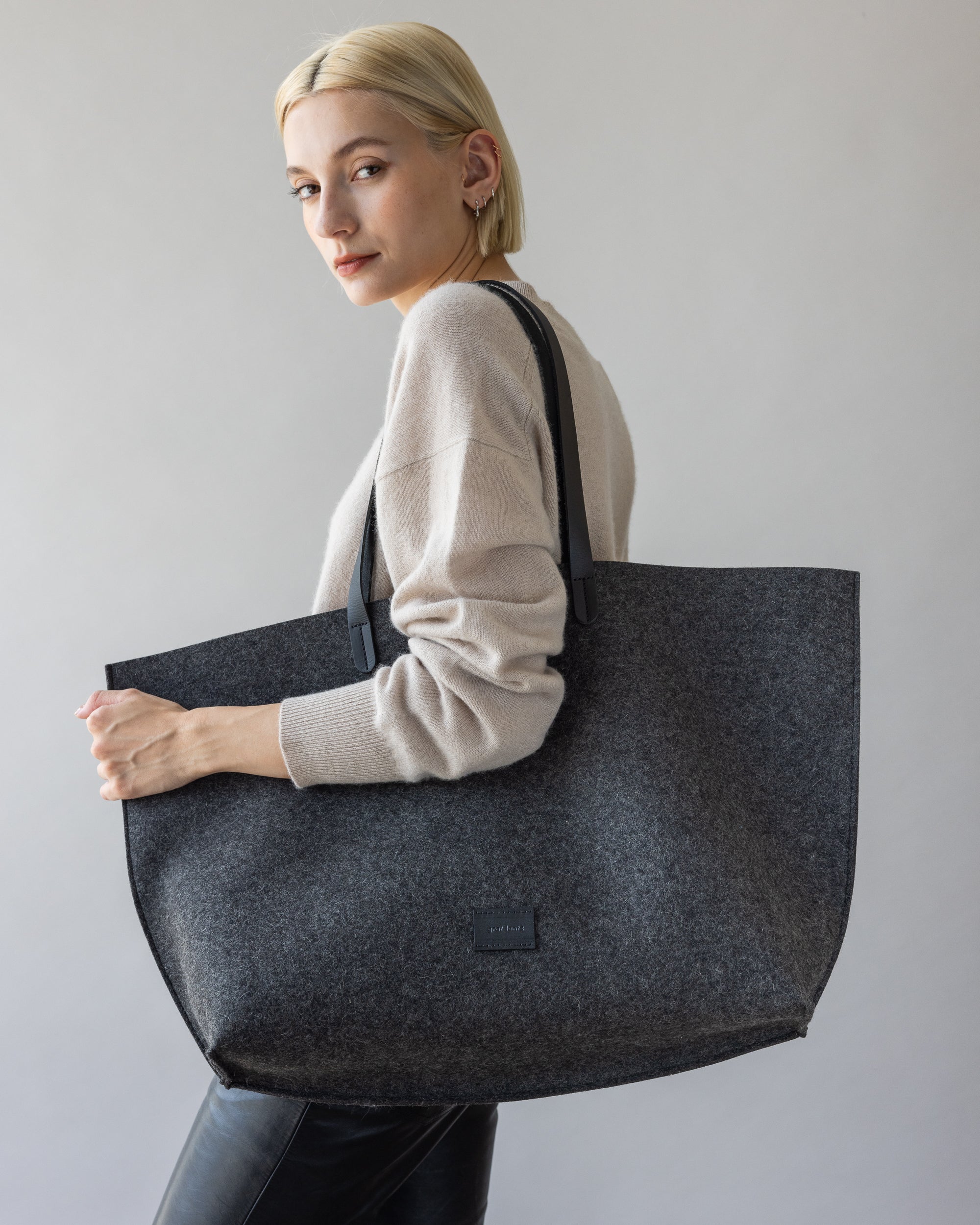 A person with short blonde hair poses with a graf lantz large gray felt tote bag with leather handles over their shoulder, wearing a sustainable Merino wool light-colored sweater and dark pants, set against a plain background.
