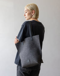 A person with short blond hair is turned slightly to the side, displaying an elegant graf lantz Hana Tote over their shoulder. Theyre wearing a black shirt and jeans against a plain, light gray background.