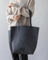 A person in a beige sweater and black pants holds the stylish Hana Tote by graf lantz, a large gray felt bag with black handles that exudes effortless style.