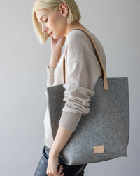 A person with short blonde hair and a neutral expression holds a Hana Tote from graf lantz, featuring leather handles. Theyre wearing a light beige Merino wool sweater and black pants, standing against a plain background.