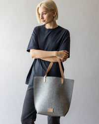 A person with short blonde hair in a black t-shirt and dark pants holds a gray Hana Tote by graf lantz, featuring beige leather handles. The background is neutral and light, creating a minimalist vibe.